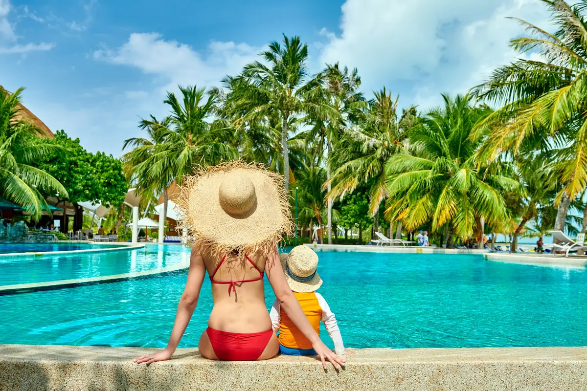 A woman and child sitting together at the edge of a palm lined pool
