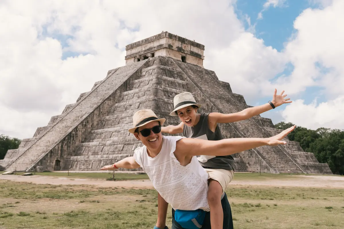 A mother carrying her daughter as they spread their arms in front of a Mayan pyramid