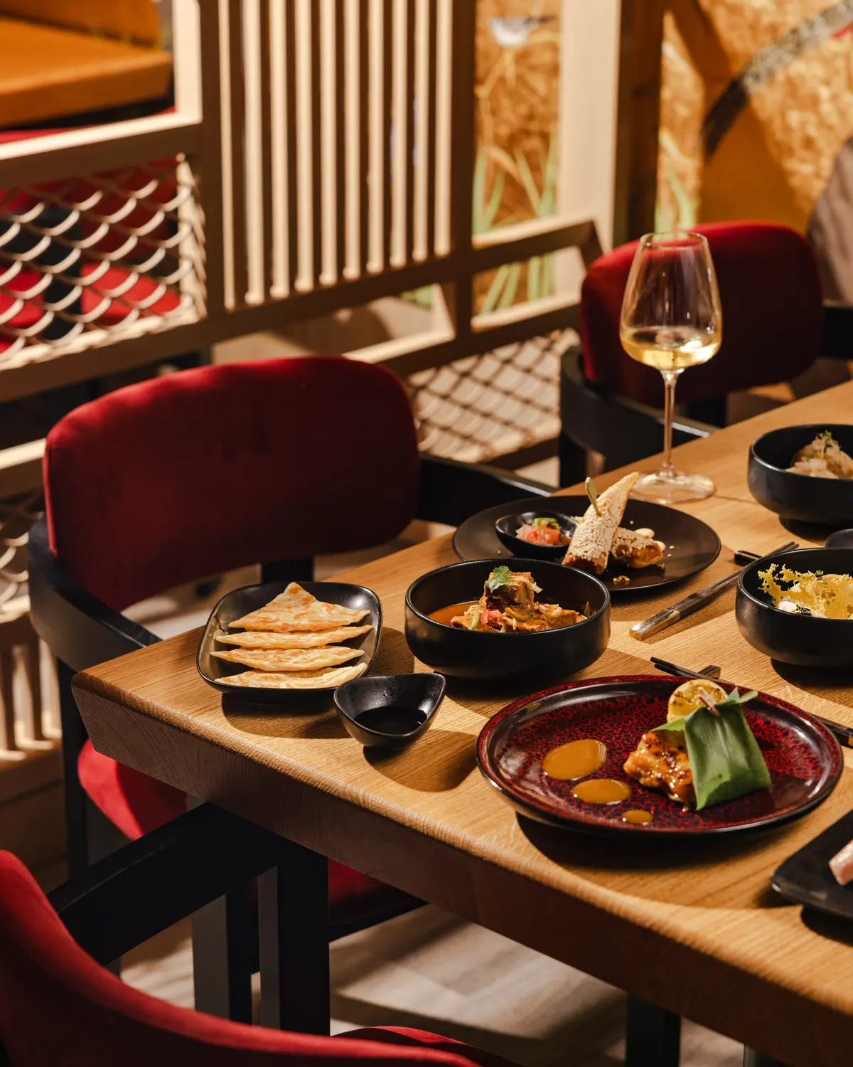 A restaurant table set with colorful dishes and wine beside red velvet chairs