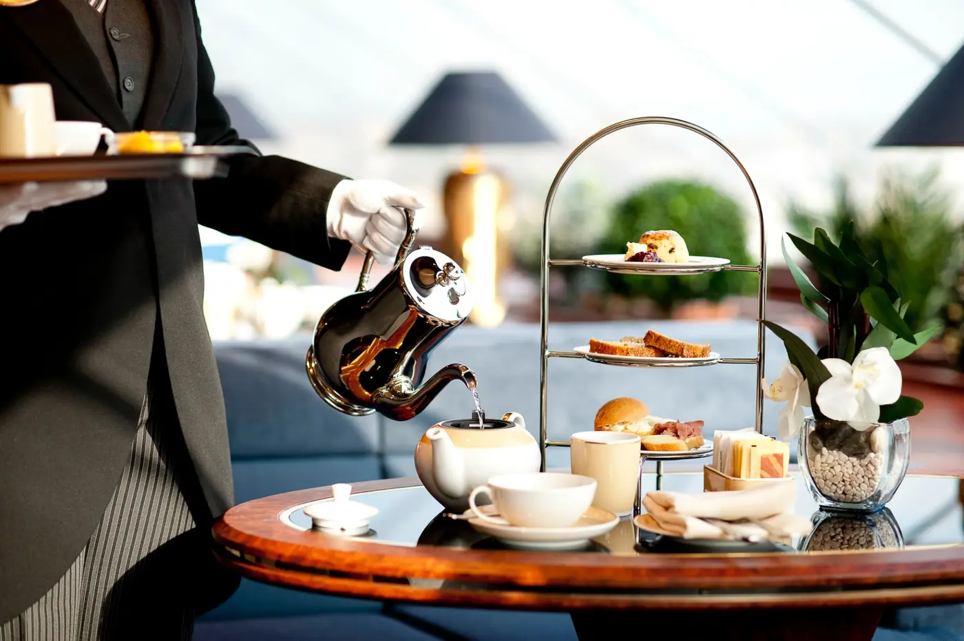 A white gloved butler pouring tea beside a tiered tray of afternoon treats