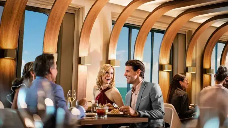Guests dining beside large ocean-view windows on a Norwegian cruise ship.