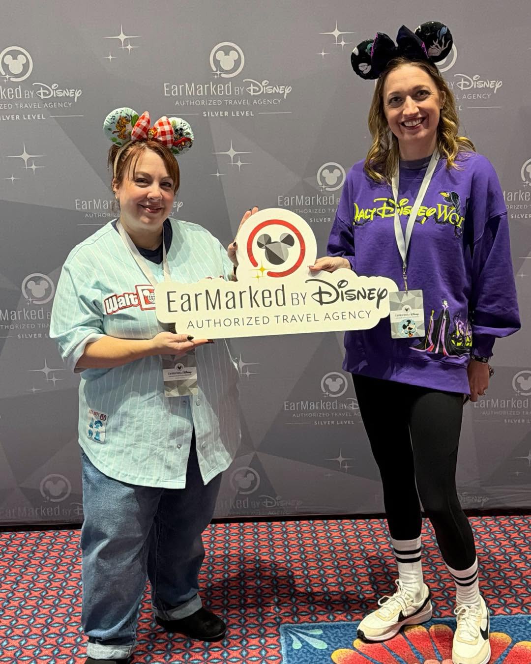 Two advisors holding an EarMarked by Disney sign in front of a Silver Level event backdrop.